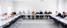 Oficjalne spotkanie grupy osób siedzących przy stołach ustawionych w podkowę w jasnej sali konferencyjnej. Wśród uczestników widoczni są przedstawiciele różnych służb mundurowych w oficjalnych mundurach (straż pożarna, wojsko, policja) oraz osoby w strojach cywilnych.