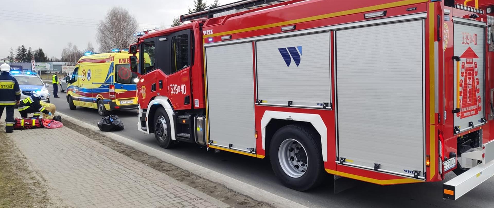 Na zdjęciu widocznych 3 strażaków w umundurowaniu specjalnym i hełmach. Obok strażaków leży torba medyczna i znajdują się oni na chodniku przy drodze krajowej. Na drodzę stoi wóz bojowy strażacki, obok karetka pogotowia i radiowóz strażacki. Na drodze ruchem kieruje umundurowany policjant. 