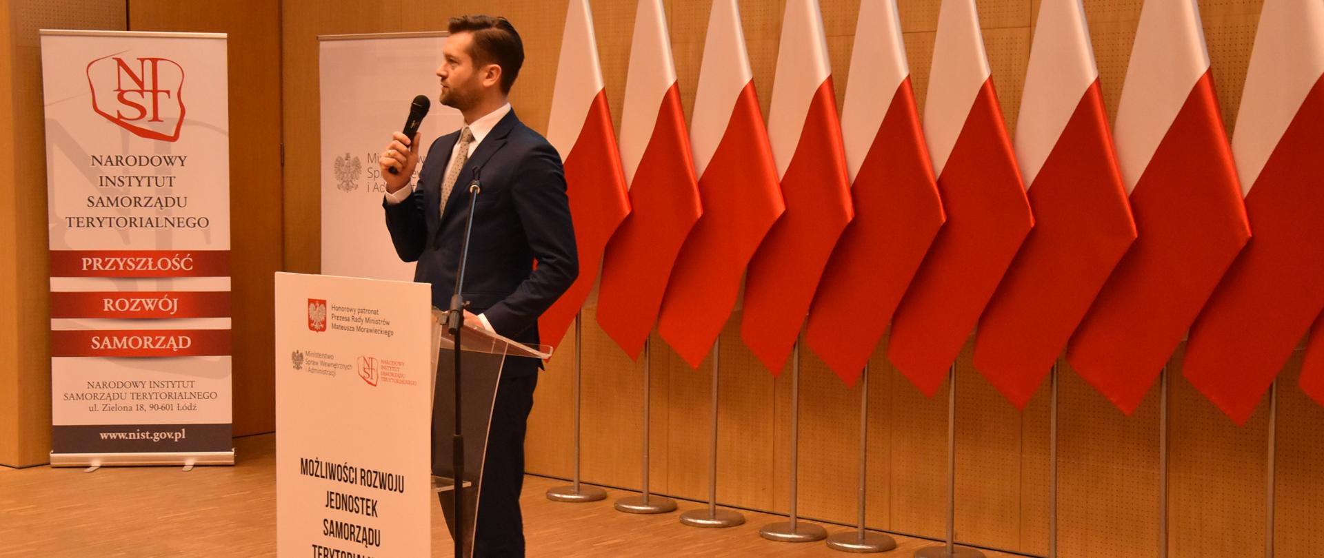 Wiceminister Kamil Bortniczuk stoi przy mównicy, w tle dziesięć biało-czerwonych flag i banery konferecyjne.