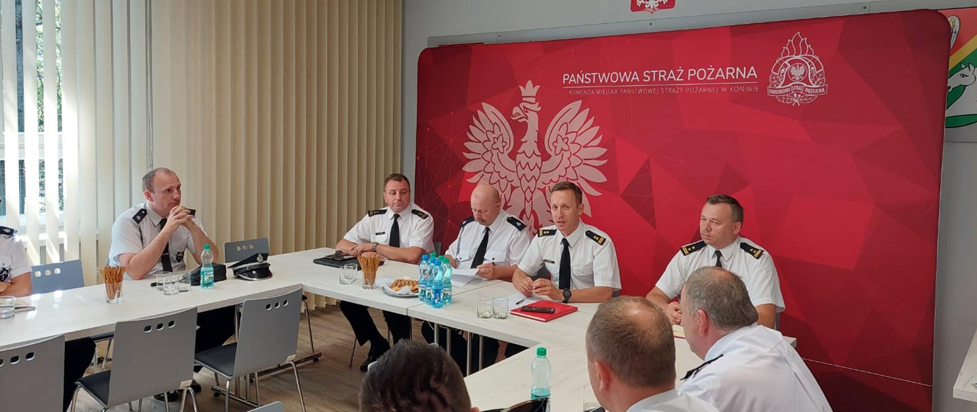 grupa strażaków w białych koszulach siedzi przy stołach, w tle duży baner z orłem