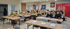 Strażacy siedzą w ławkach mają ubrane piaskowe i czarne mundury. Z tyłu widać godło Polski na ścianie. Wszystkie osoby znajdują się na sali w której rozmieszczone są ławki przy których siedzą strażacy biorący udział w szkoleniu