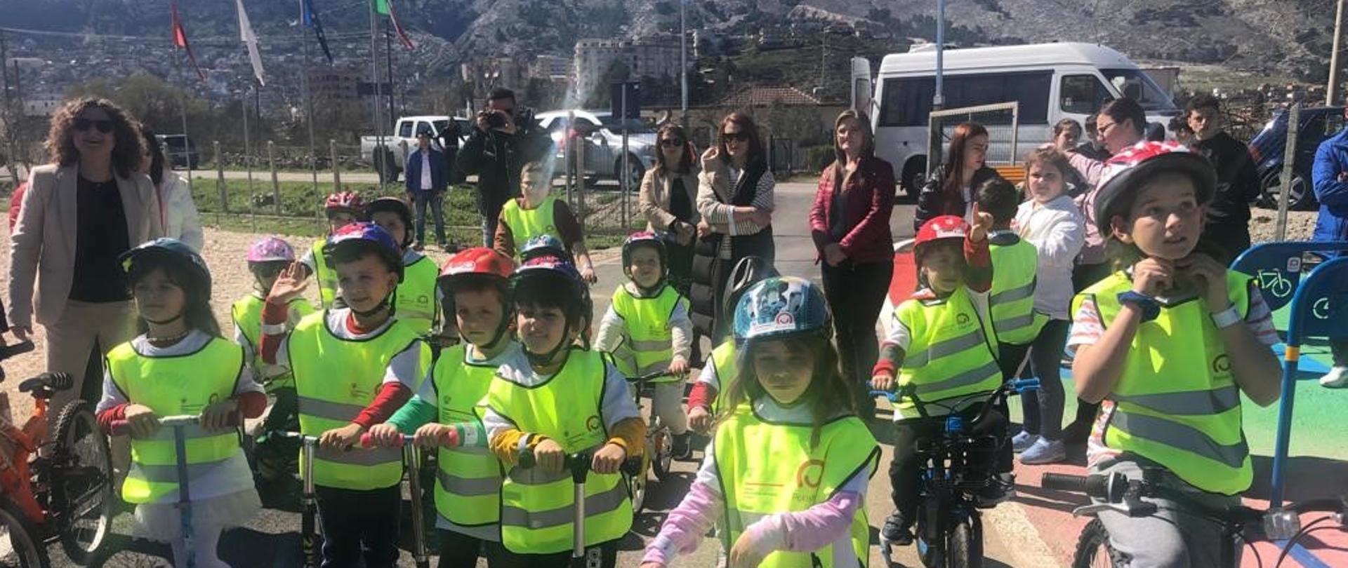 Widok z przodu. Dzieci ubrane w żółte, odblaskowe kamizelki i kaski rowerowe. Dzieci w pierwszym rzędzie stoją na hulajnogach. Dzieci w drugim rzędzie siedzą na rowerach. Za dziećmi stoją dorośli ludzie. W tle widać drogę z zaparkowanymi samochodami i góry. 