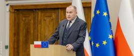 Mężczyzna w szarym garniturze przemawia z mównicy. Z przodu mównicy małe flagi Polski i Unii Europejskiej.