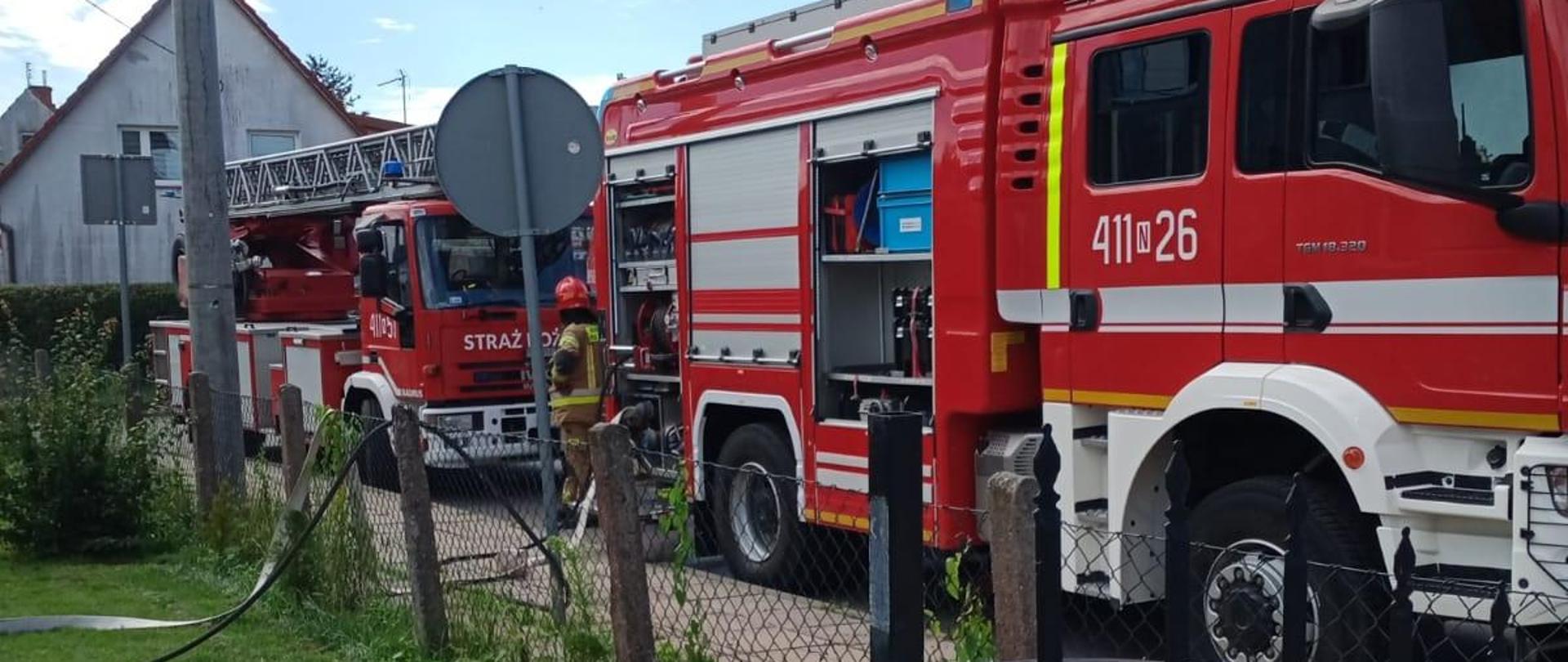 Na zdjęciu przy chodniku stoją dwa samochody strażackie. Niebieskie niebo, pogodnie.