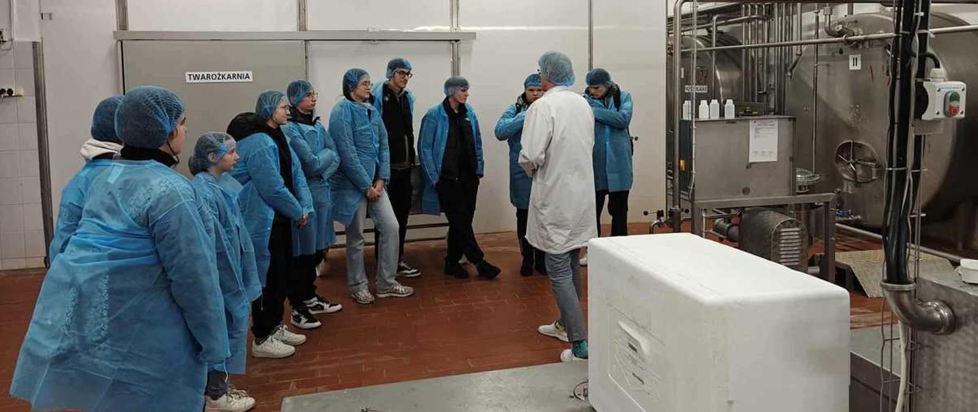 Zdjęcie przedstawia grupę osób na wycieczce w zakładzie produkcyjnym (twarożkarni).
Wszyscy uczestnicy mają na sobie odzież ochronną (niebieskie fartuchy i siatki na włosy). Stoją w rzędzie, obserwując prowadzącego (mężczyzna w białym kitlu), który stoi tyłem do nich. W tle widoczne są maszyny przemysłowe ze stali nierdzewnej, a na ścianie znajduje się napis "TWAROŻKARNIA".