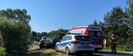 Na pierwszym planie radiowóz policyjny, obok niego karetka pogotowia ratunkowego. W tle spalony samochód osobowy, który brał udział w wypadku i ma uszkodzony cały przód pojazdu. Obok karetki pogotowia stoi strażak, który wcześniej prowadził działania gaśnicze. 