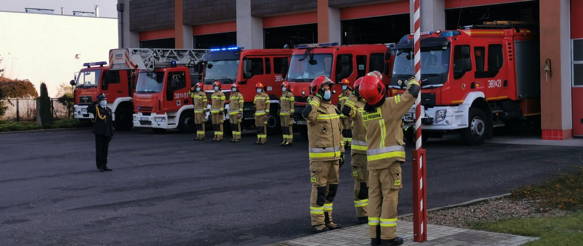 Strażacy wciągają flagę na maszt. W tle stoją strażacy w szeregu i samochody strażackie.