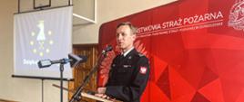 Brygadier Tomasz Grelak przemawia z mównicy w trakcie narady rocznej. W tle baner Państwowej Straży Pożarnej i ekran na którym prezentowana jest statystyka podsumowująca działania.