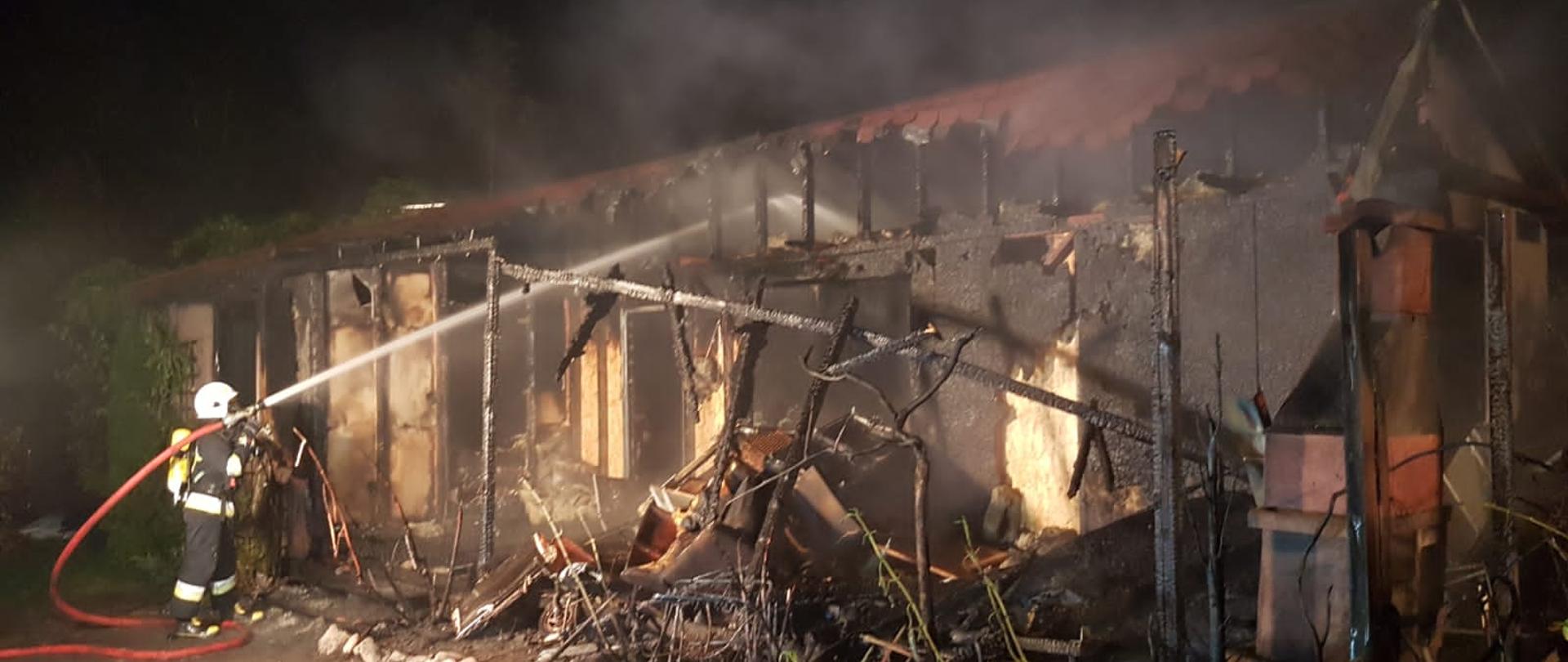 Domek letniskowy całkowicie spalony. Na pierwszym planie strażak dogaszający pogorzelisko.