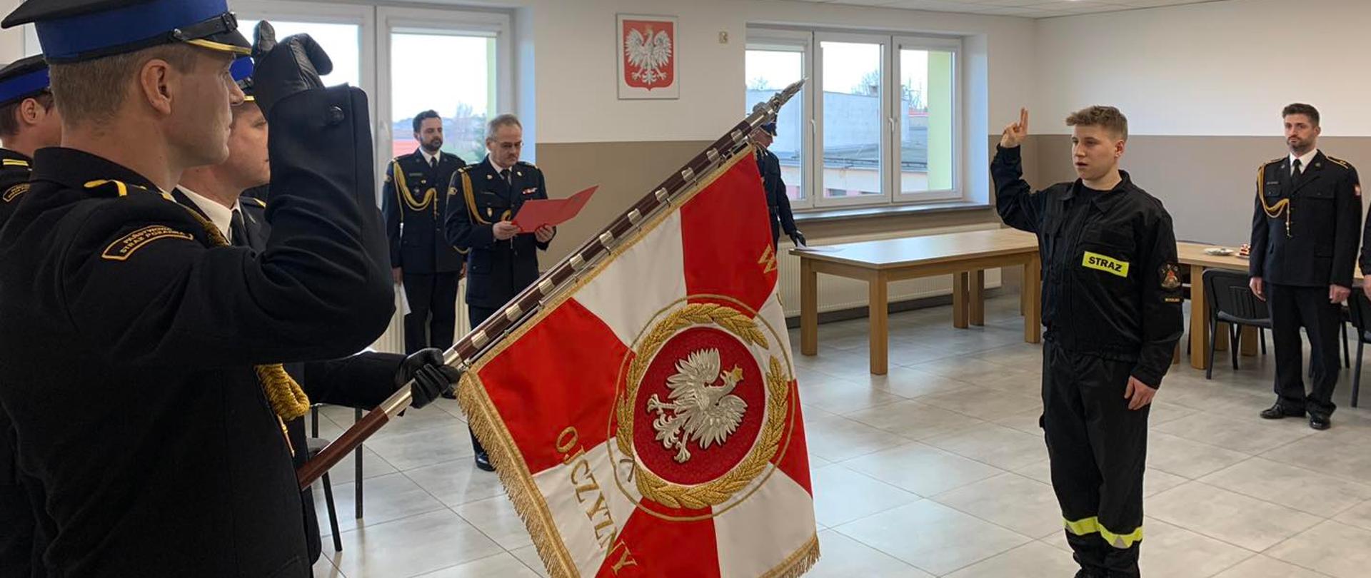Nowo przyjęty strażak składa rotę ślubowania. Poczet sztandarowy oddane honor poprzez opuszczenie sztandaru i salutowanie. 