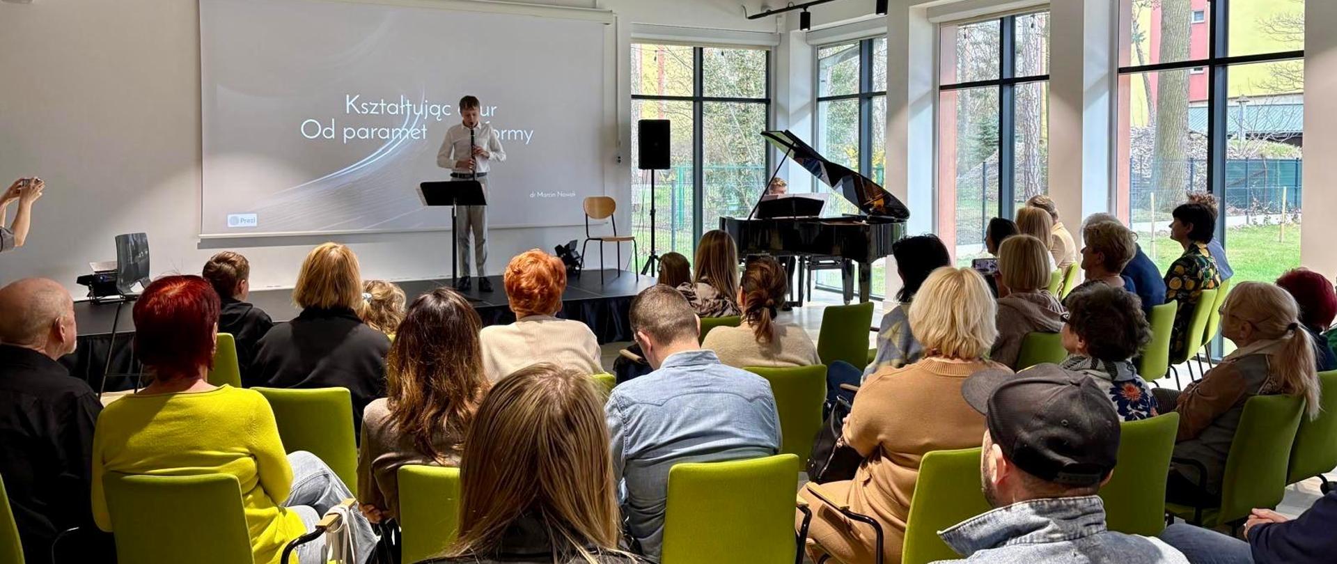 Widok sali konferencyjnej, w której odbywa się koncert. Na scenie stoi mężczyzna grający na klarnecie, a obok niego, przy czarnym pianinie, siedzi pianista. Za nimi znajduje się ekran z napisem 'Kształtując ażurową formę: Od parametru do formy'. W sali siedzi kilka osób, w tym dzieci, które obserwują występ. Sala jest nowocześnie urządzona, z dużymi oknami wpuszczającymi naturalne światło.