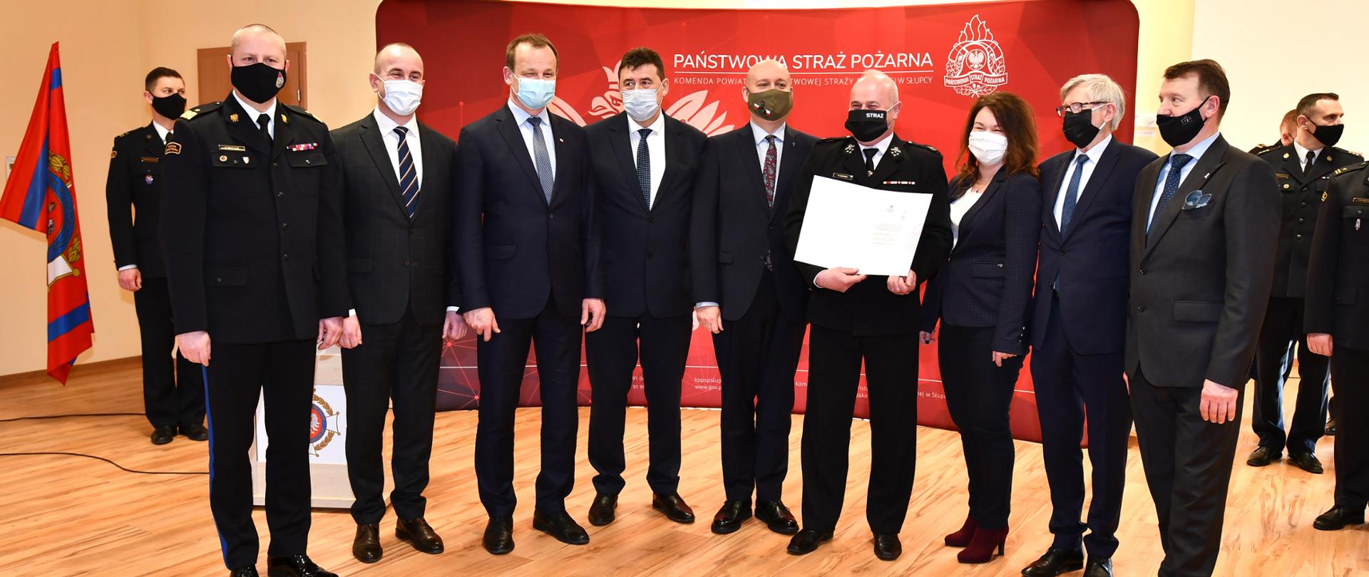 Przedstawiciel jednostki OSP z otrzymana promesą stoi wraz z nim sześc osób w garniturach jedna kobieta i funkcjonariusz