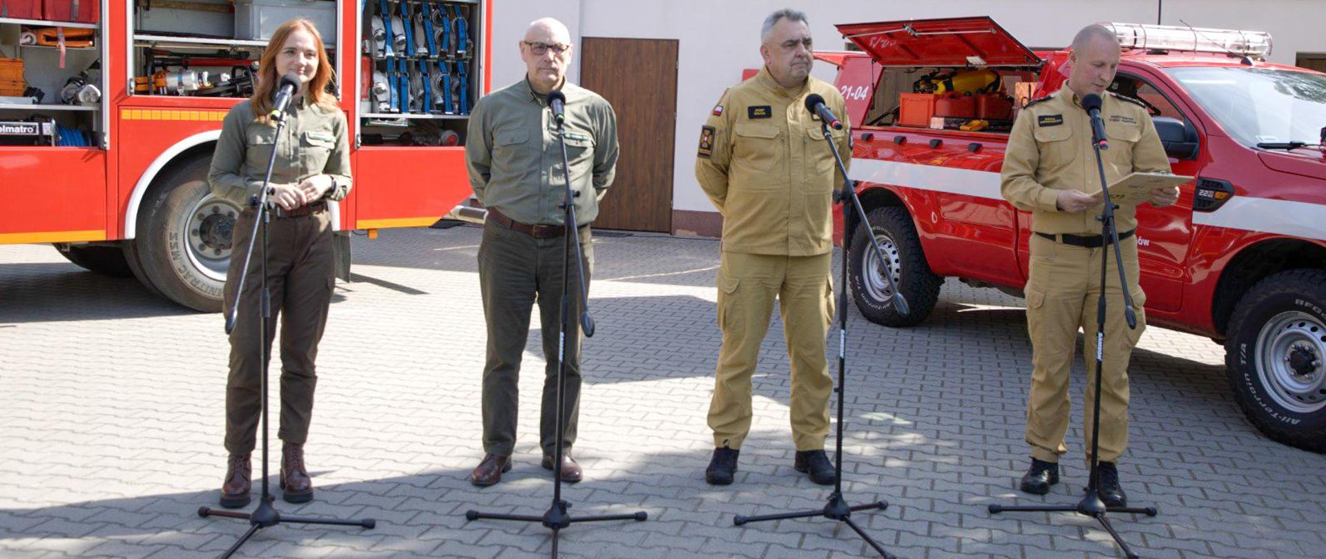 Przedstawiciele służb ratowniczych i leśnych przemawiający przed mikrofonami, na placu przed budynkiem i samochodami strażackimi. W tle widoczna baza sprzętu pożarniczego.
