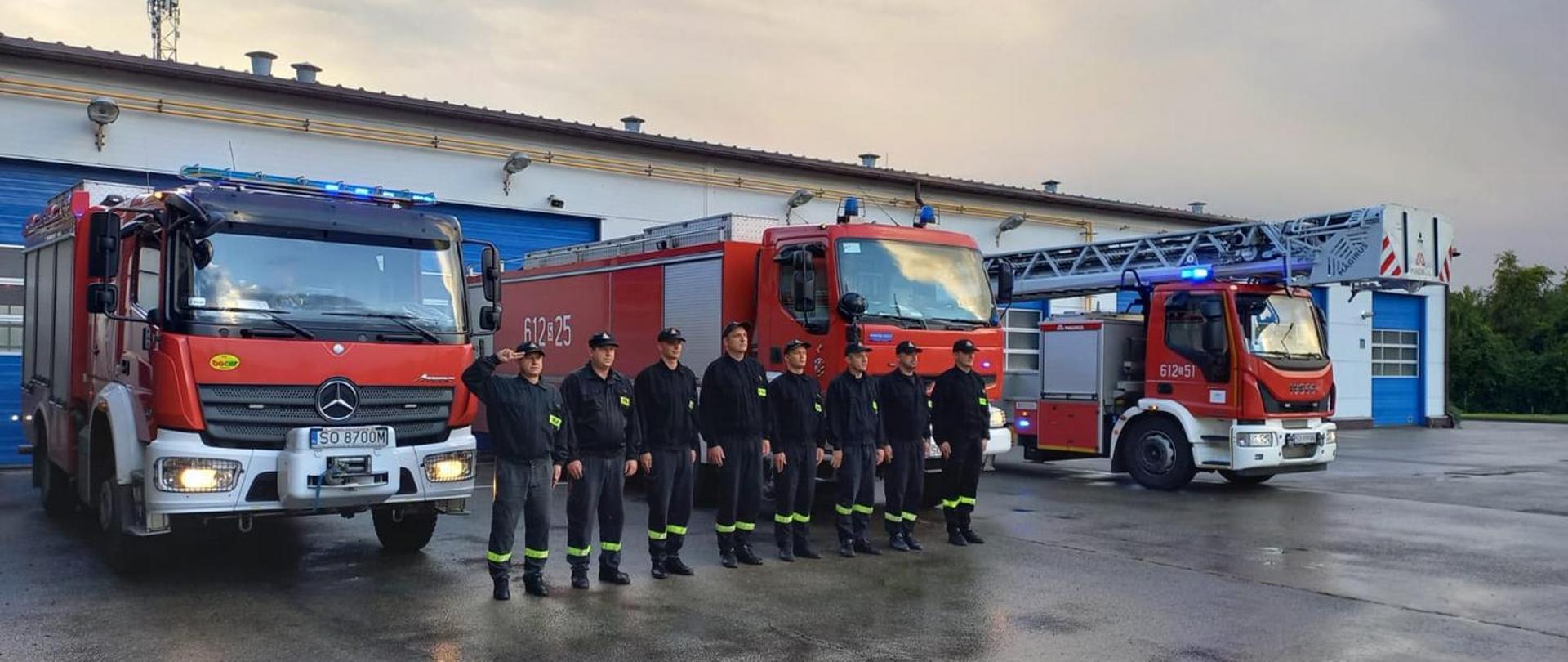 Strażacy Komendy Miejskiej PSP w Sosnowcu ustawieni w szeregu w umundurowaniu koszarowym przed samochodami ratowniczo-gaśniczymi z włączoną sygnalizacją świetlną. Strażacy oddają hołd zmarłemu strażakowi.