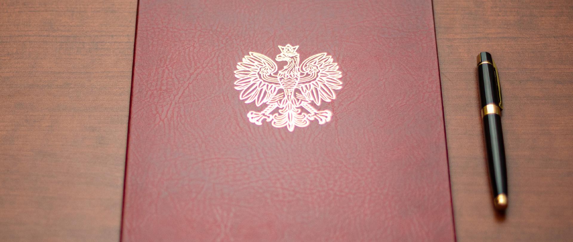 Na zdjęciu widać twardą oprawę na dokumenty z wytłoczonym orłem z godła narodowego i leżące obok pióro wieczne.