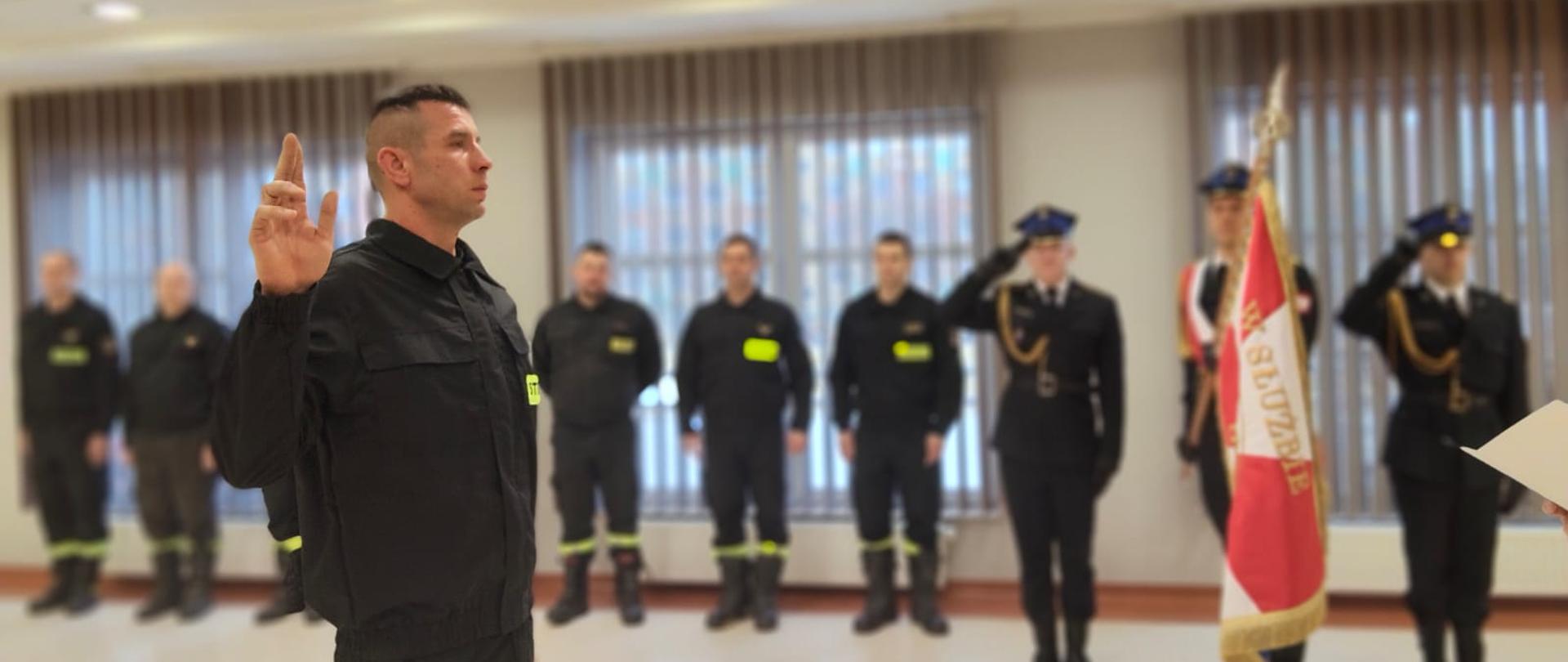Na zdjęciu widoczny strażak w postawie do ślubowania. W tle strażacy uczestniczący w ceremonii oraz sztandar Komendy Powiatowej PSP.