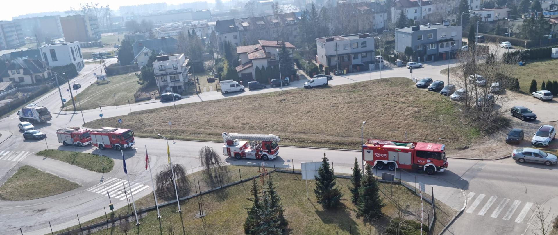 Widok z lotu ptaka na ulicę dojazdową do szpitala, cztery samochody pożarnicze wjeżdżają na teren ćwiczeń, w oddali budynki mieszkalne, drogi.