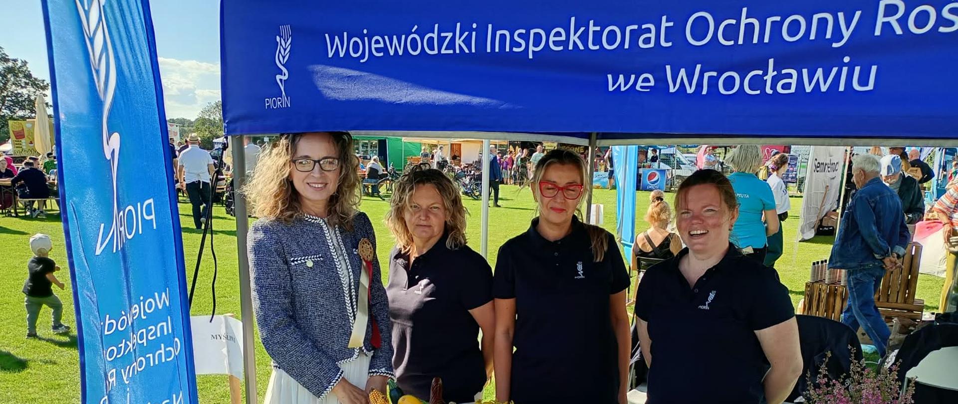 Na plenerowym stoisku Wojewódzkiego Inspektoratu Ochrony Roślin i Nasiennictwa we Wrocławiu stoją cztery kobiety. Na stole przed nimi znajdują się kosze z warzywami, rękodzieło, materiały informacyjne i gadżety promocyjne. Stoisko jest oznaczone niebieskimi banerami z nazwą inspektoratu. W tle widać uczestników plenerowego wydarzenia na terenie zielonym.