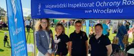 Na plenerowym stoisku Wojewódzkiego Inspektoratu Ochrony Roślin i Nasiennictwa we Wrocławiu stoją cztery kobiety. Na stole przed nimi znajdują się kosze z warzywami, rękodzieło, materiały informacyjne i gadżety promocyjne. Stoisko jest oznaczone niebieskimi banerami z nazwą inspektoratu. W tle widać uczestników plenerowego wydarzenia na terenie zielonym.