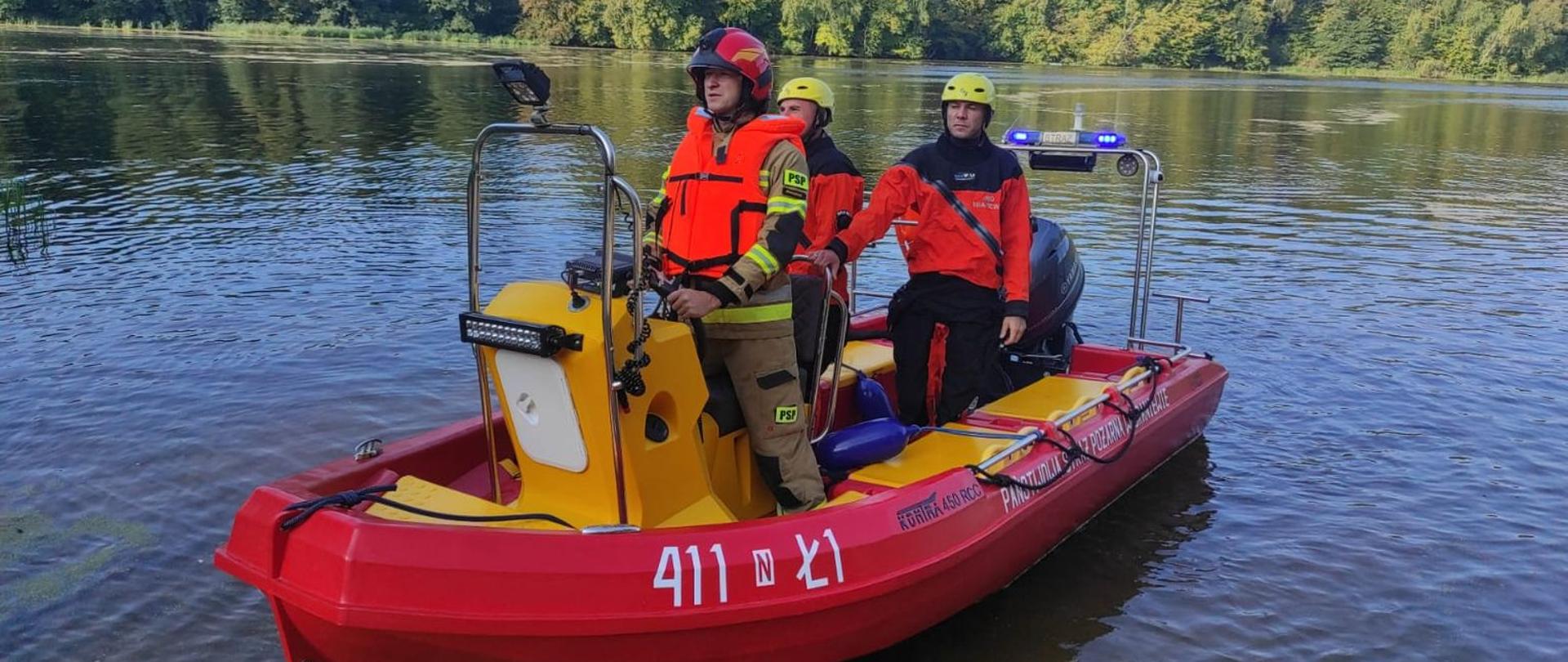 Na jeziorze stoi czerwona lódź strażacka a na niej trzech strażaków ubranych w kombinezony do pracy w wodzie.