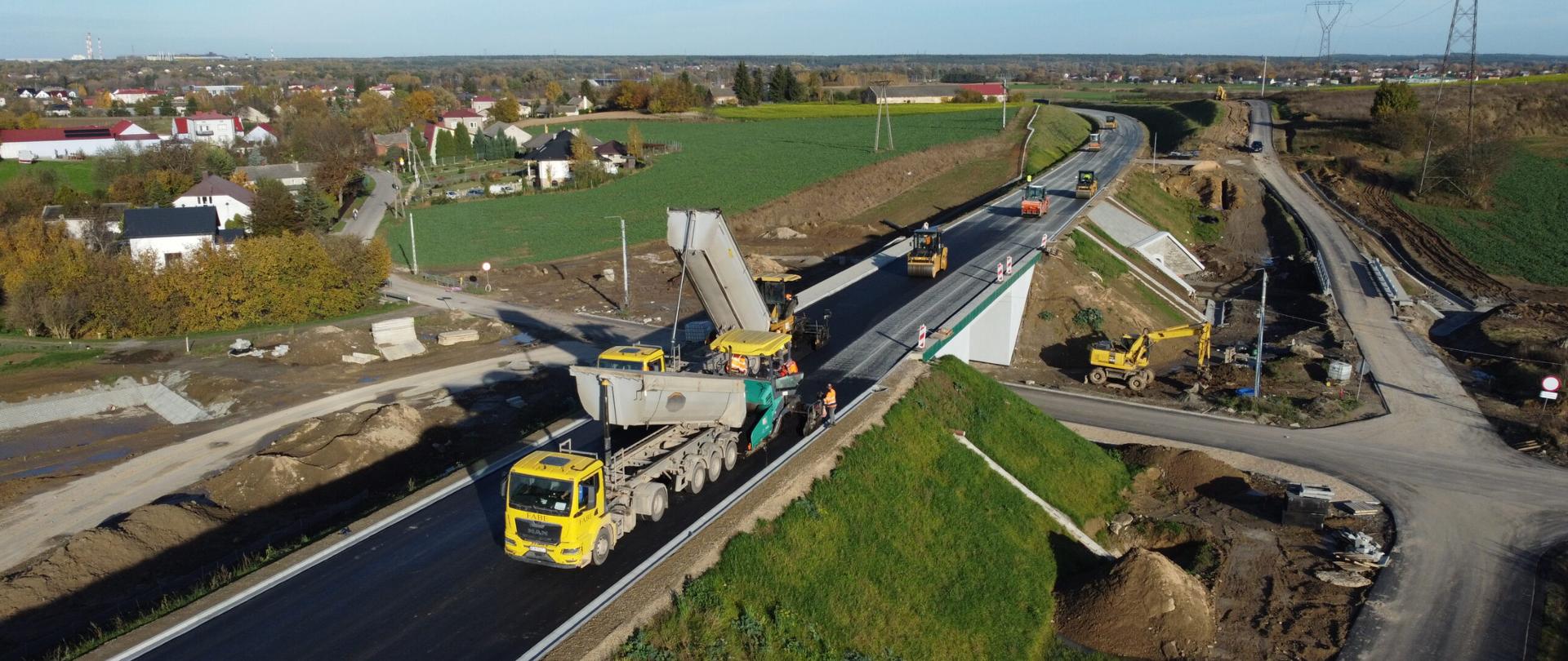 DK9 obwodnica Ostrowca Świętokrzyskiego w budowie - układanie nawierzchni asfaltowej (czarnej) na jednojezdniowej drodze. Na budowanej trasie samochody ciężarowe, rozściełacze i walce drogowe. Na skrzyżowaniu obwodnicy z drogą lokalną wiadukt. Pod wiaduktem koparka. W oddali domy jednorodzinne i zielone pola