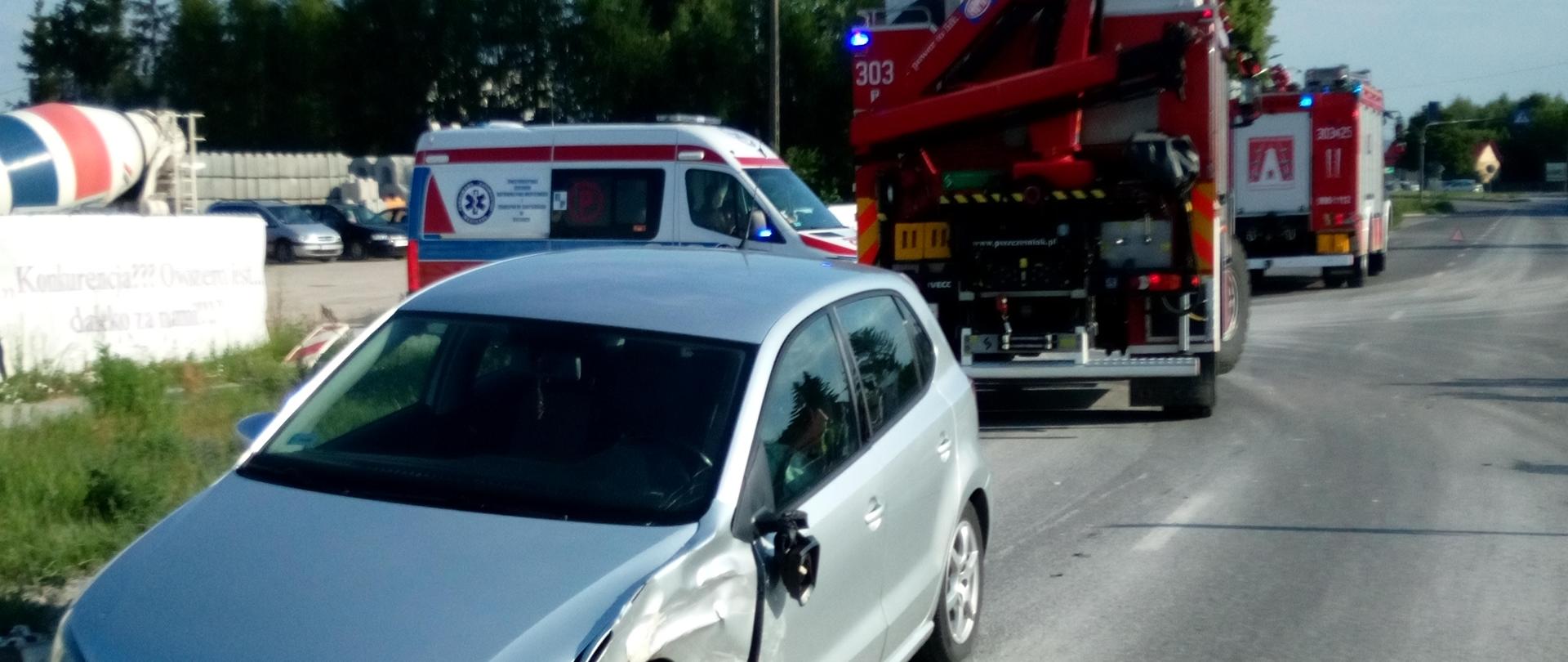 Zdjęcie przedstawia uszkodzony pojazd osobowy - w tle widać Pogotowie Ratunkowe oraz samochody Straży Pożarnej. 