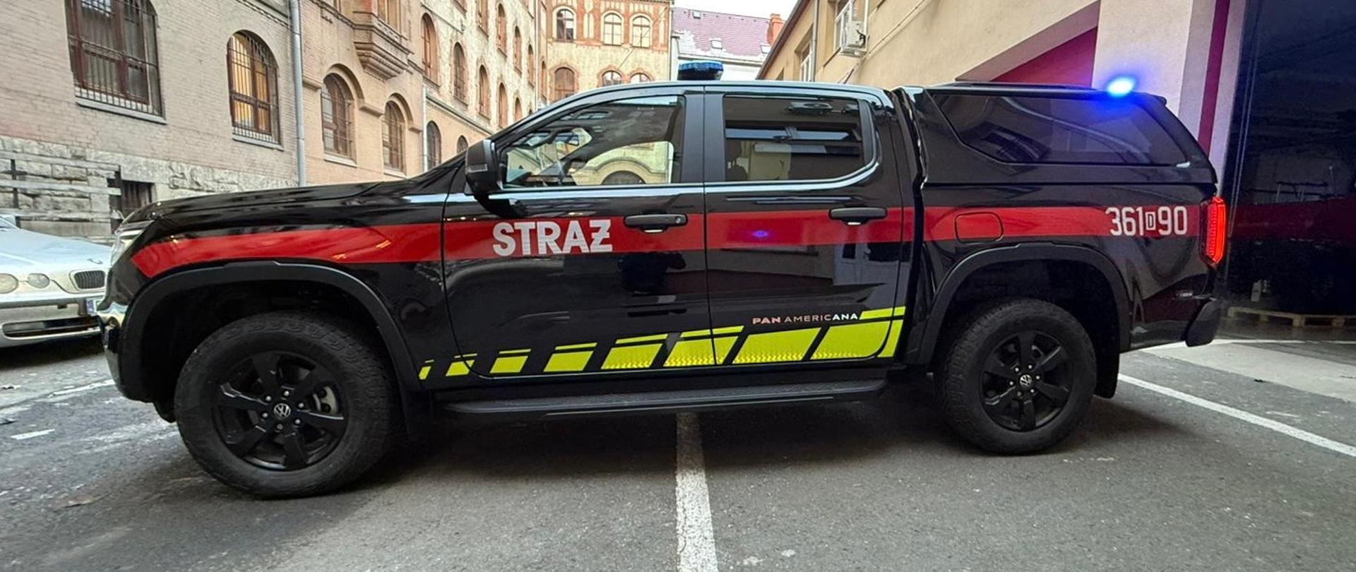 Zdjęcie przedstawia nowoczesny, czarny samochód terenowy typu pickup marki Volkswagen Amarok, przystosowany do celów ratowniczych i zaparkowany w garażu straży pożarnej. Pojazd posiada charakterystyczne oznakowanie w postaci szerokiego czerwonego pasa wzdłuż nadwozia z białym napisem „STRAŻ” na drzwiach oraz odblaskowe, żółto-czarne pasy u dołu i na masce. Na dachu samochodu zamontowana jest belka z niebieskimi światłami ostrzegawczymi i napisem „STRAŻ”, a na przednim grillu widnieje logo marki oraz oznaczenie silnika V6. W tle widoczne jest profesjonalne zaplecze jednostki: czerwone metalowe szafki z zawieszonymi mundurami bojowymi, ułożone w rzędzie czerwone hełmy strażackie oraz system odciągu spalin. Całość zdjęcia, wykonanego w techniczny i przejrzysty sposób, prezentuje gotowość bojową oraz nowoczesne wyposażenie formacji.