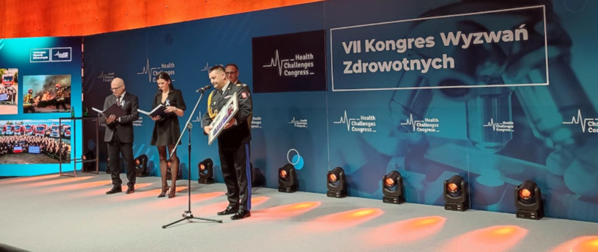 Na scenie dwóch prowadzących galę - kobieta i mężczyzna oraz dwie osoby wyróżnione. Przed mikrofonem odbierający w imieniu Komendanta Głównego PSP nadbryg. Andrzeja Bartkowiaka - Śląski Komendant Wojewódzki PSP nadbryg. Jacek Kleszczewski, w tle napis VII Kongres Wyzwań Zdrowotnych