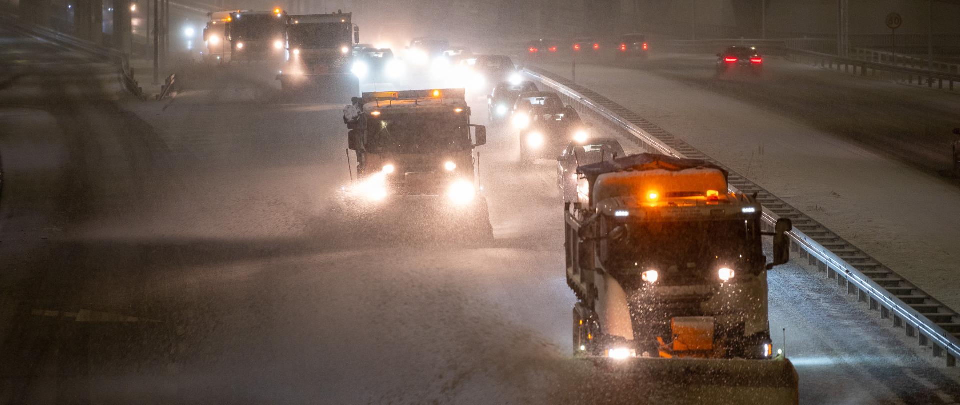 Pługosolarki jadące po zaśnieżonej drodze ekspresowej nocną porą. Za nimi podążają samochody osobowe. Pada śnieg. 