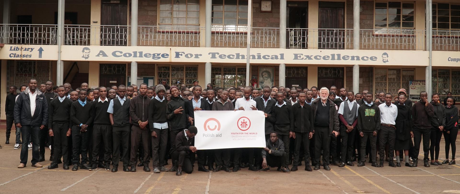 Grupa ok. 100 osób pozuje do zdjęcia przed szkołą trzymając przed sobą plakat z logo polskiej pomocy i SWM