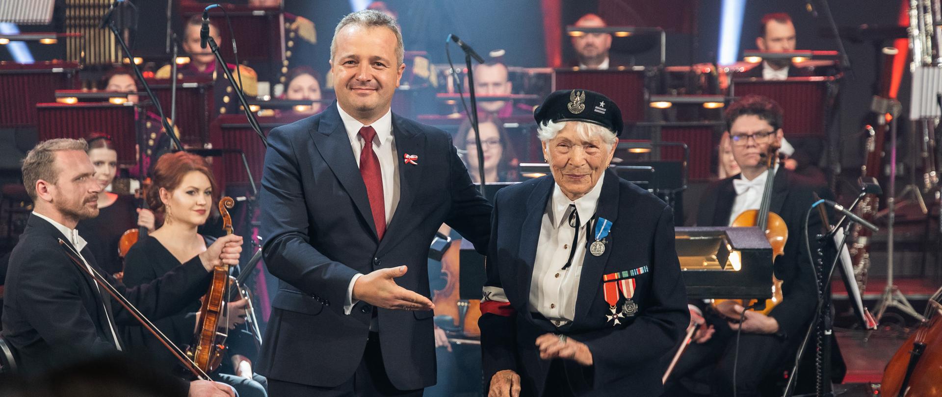 dwie osoby na scenie, w tle muzycy - wojewoda i odznaczona