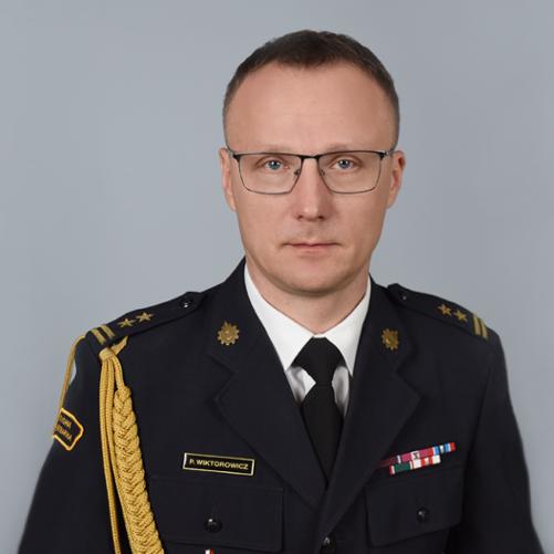 Zdjęcie to oficjalny portret mężczyzny w mundurze wyjściowym Państwowej Straży Pożarnej, przedstawiony na jednolitym, szarym tle.
Postać
• Mężczyzna ma krótkie, ciemne włosy, nosi okulary w ciemnych oprawkach i patrzy bezpośrednio w stronę obiektywu z poważnym wyrazem twarzy.
• Jest ubrany w ciemnogranatowy mundur wyjściowy, białą koszulę oraz czarny krawat.
Detale munduru
Mundur jest bogato zdobiony insygniami świadczącymi o stopniu i zasługach:
• Akselbanty: Na prawym ramieniu widoczny jest gruby, pleciony sznur galowy (oficerski) w kolorze złotym.
• Pagony: Na ramionach znajdują się pagony z dwiema złotymi gwiazdkami i dwiema belkami, co odpowiada stopniowi brygadiera.
• Baretki: Nad lewą kieszenią przypięte są baretki odznaczeń państwowych i resortowych w różnych kolorach.
• Tabliczka imienna: Nad prawą kieszenią znajduje się czarna plakietka ze złotym napisem: P. WIKTOROWICZ.
• Emblematy: Na kołnierzu widoczne są złote emblematy korpusu straży pożarnej (ogniki). Na prawym rękawie znajduje się częściowo widoczna naszywka z napisem „PAŃSTWOWA STRAŻ POŻARNA”.
Zdjęcie charakteryzuje się profesjonalnym oświetleniem, jest ostre i ma wyważoną kompozycję typu popiersie.
