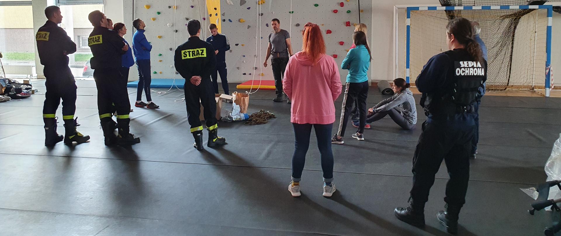 Na zdjęciu widać strażaków w ubraniach koszarowych oraz nauczycieli z ZSP nr1 którzy biorą udział w szkoleniu z wspinaczki na ściance. Ścianka znajduje się za wszystkimi osobami.