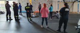 Na zdjęciu widać strażaków w ubraniach koszarowych oraz nauczycieli z ZSP nr1 którzy biorą udział w szkoleniu z wspinaczki na ściance. Ścianka znajduje się za wszystkimi osobami.