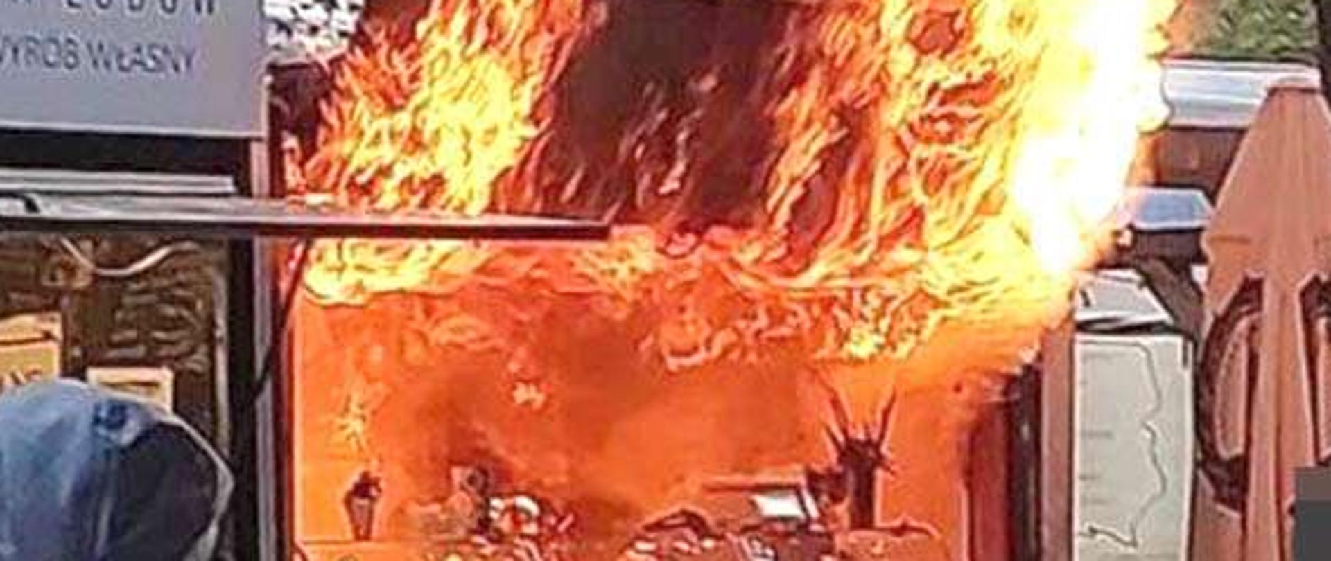 Duże płomienie wydobywające się z wnętrza pawilonu gastronomicznego. Na przodzie stoiska napisy "kurtosz" i "kołacz"