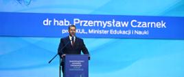 Na tle niebieskiej ściany z napisem Dr hab. Przemysław Czarnek stoi za mównicą minister Czarnek i mówi do mikrofonu, z przodu na podłodze leży bukiet białych kwiatów.