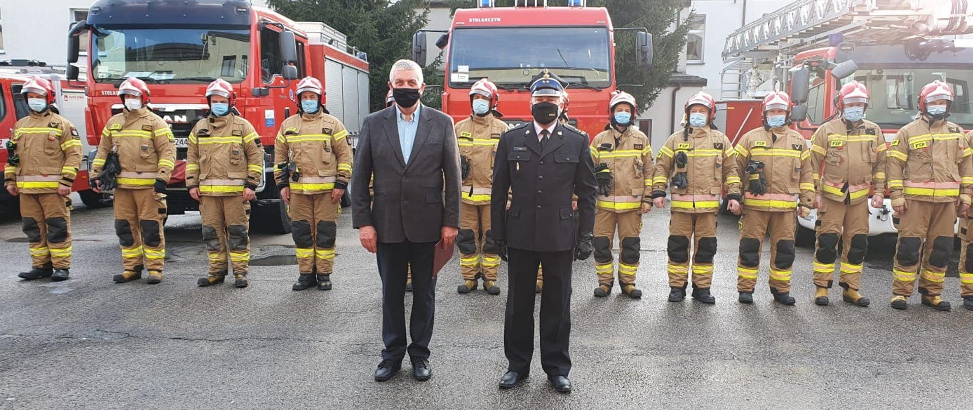 Komendant Powiatowy w mundurze wyjściowym oraz pracownik cywilny stoją przed funkcjonariuszami w ubraniach specjalnych ustawionych w szeregu. W tle widać samochody strażackie.
