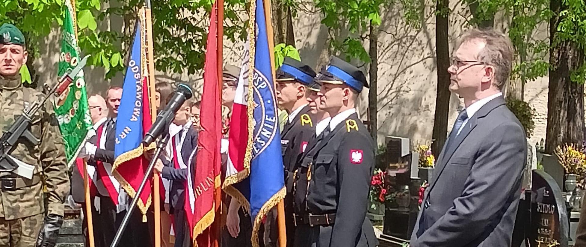 Na cmentarzu widoczne poczty sztandarowe służb mundurowych widoczny żołnierz oraz osoba cywilna