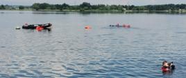 Na zdjęciu widoczny akwen. W oddali drzewa. W wodzie znajduje się łódź pontonowa. Obok pływa czterech strażaków w czerwonych, suchych skafandrach nurkowych.