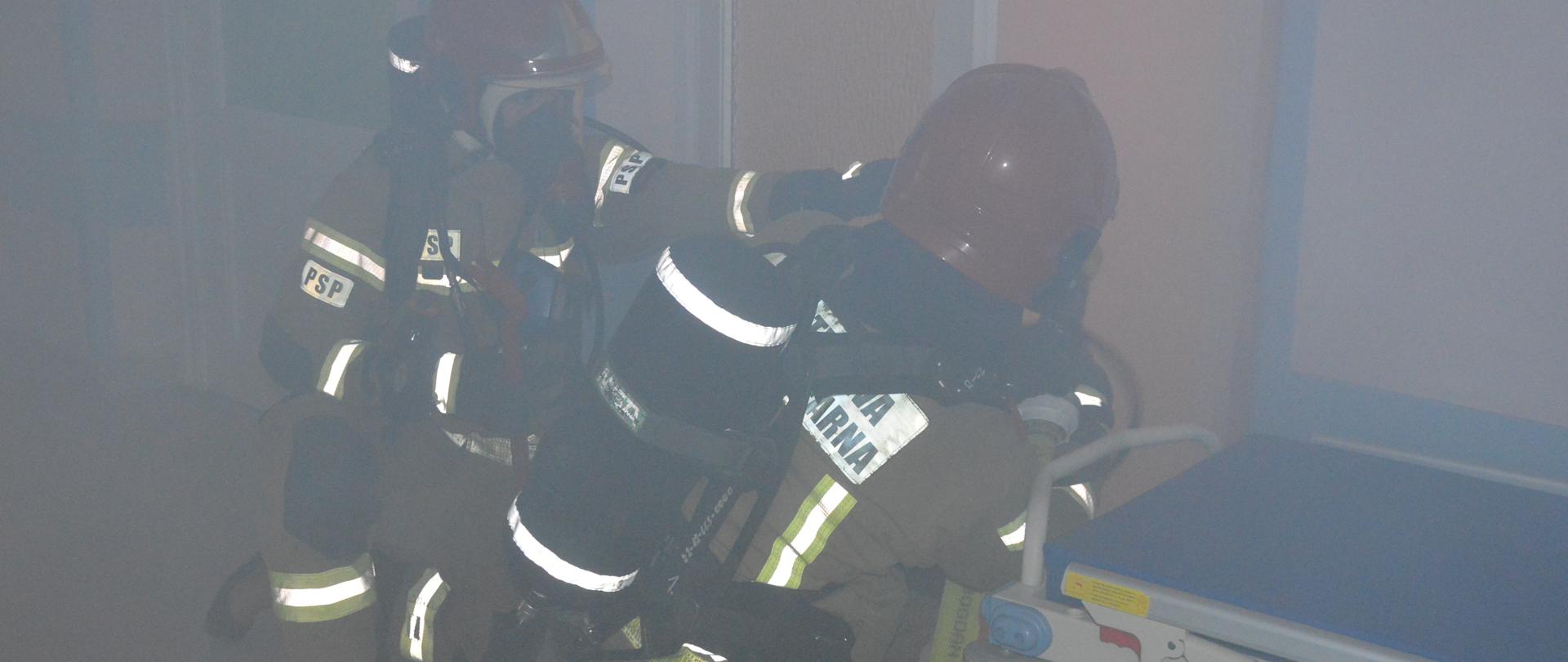 Ćwiczenia w szpitalu. Jedna z sal na oddziale chirurgicznym. W pomieszczeniu, które jest zadymione znajduje się dwóch strażaków. Mają czerwone hełmy, a na plecach założony aparat powietrzny. Pierwszy z nich trzyma w rękach wąż z podłączoną prądownicą. Obaj są w pozycji klęczącej.