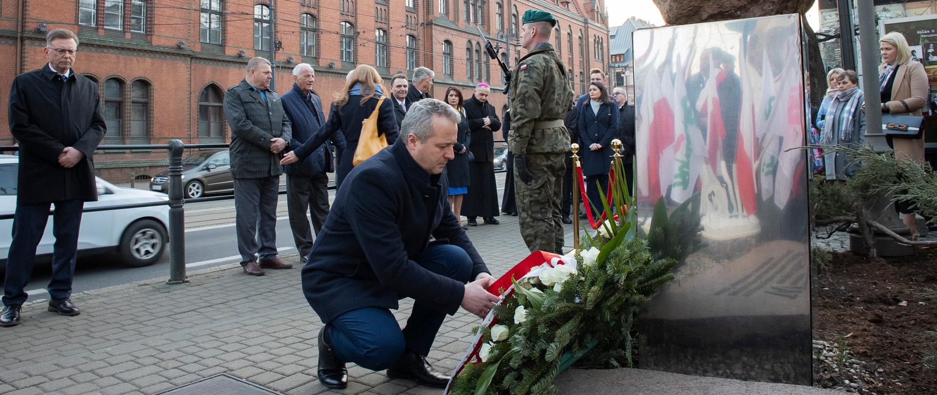 Ludzie stojący przed pomnikiem i 1 osoba składająca kwiaty