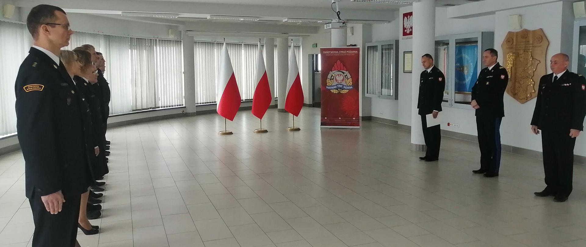 Po prawej stronie trzech mężczyzn ubranych w czarne mundury wyjściowe i białe koszule stoją na Sali konferencyjnej KP PSP w Bochni. Po lewej stornie kilkunastu strażaków ubranych w czarne mundury wyjściowe oraz ubrania koszarowe stoją na baczność. W tle na ścianie sztandary w gablotach szklanych, flagi koloru biało czerwonego, żaluzje koloru szarego