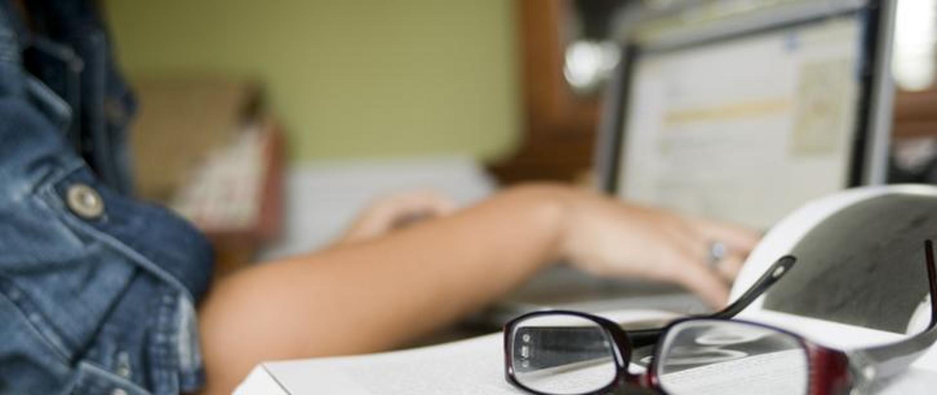 Obraz z osobą przy komputerze i leżącymi okularami.