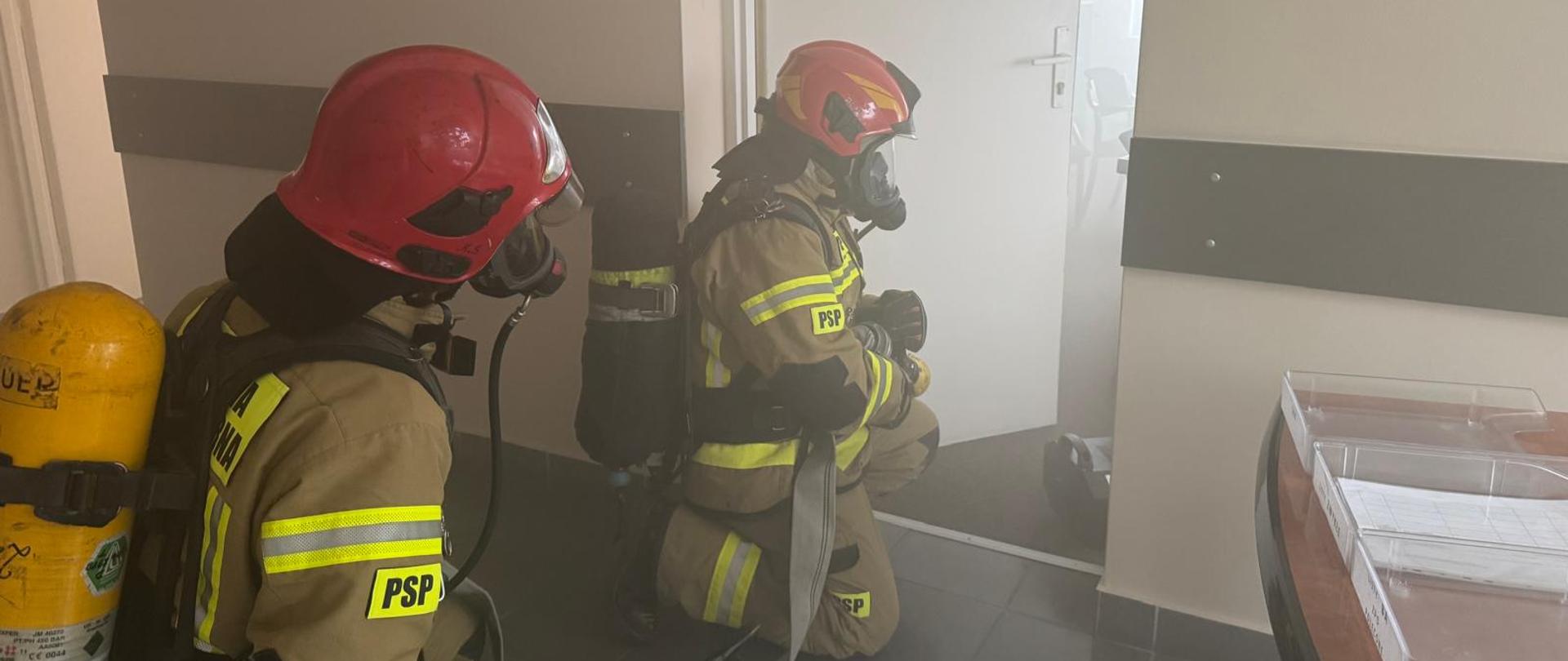 Na zdjęciu widać dwóch strażaków w pełnym ekwipunku, którzy pracują wewnątrz urzędu. Jeden z nich trzyma wąż gaśniczy, drugi podąża za nim. Strażacy rozwijają wąż strażacki, mają ubrany sprzęt ochrony układu dróg oddechowych, oraz przygotowują się do akcji.