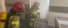 Na zdjęciu widać dwóch strażaków w pełnym ekwipunku, którzy pracują wewnątrz urzędu. Jeden z nich trzyma wąż gaśniczy, drugi podąża za nim. Strażacy rozwijają wąż strażacki, mają ubrany sprzęt ochrony układu dróg oddechowych, oraz przygotowują się do akcji.