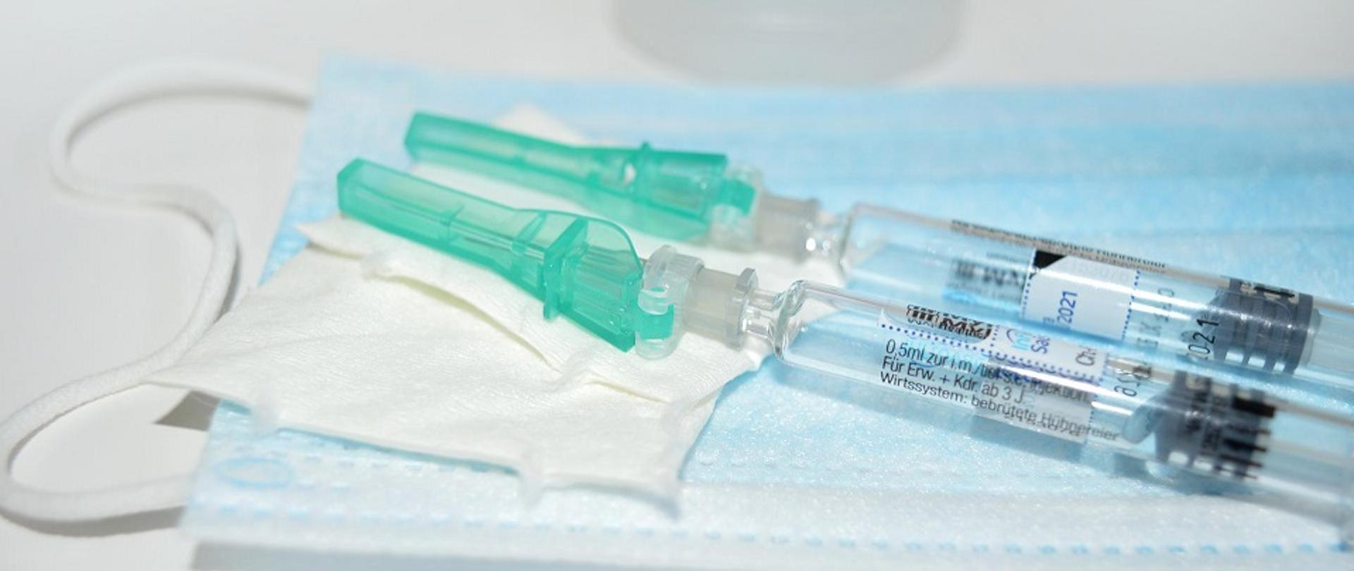 Dwie strzykawki z igłami ze szczepionkami leżące na wacikach i na maseczce ochronnej jednorazowej.
