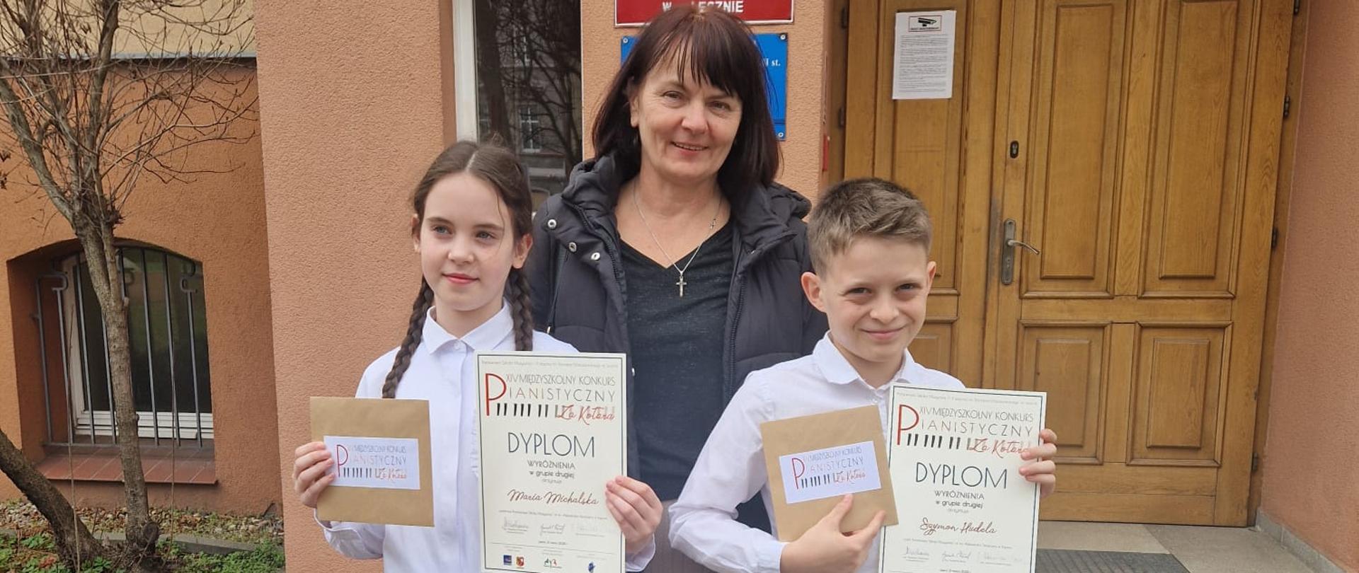 Na zdjęciu przed wejściem do budynku szkoły muzycznej stoją trzy osoby – nauczycielka w środku oraz dwoje uczniów (chłopiec i dziewczynka) po jej bokach. Dzieci trzymają w rękach dyplomy i koperty. Dziewczynka z lewej ma długie, ciemne włosy splecione w warkocze, białą bluzkę, czarną spódniczkę i białe rajstopy w kropki. Chłopiec z prawej ma krótkie jasne włosy, białą koszulę i granatowe spodnie. Nauczycielka pośrodku nosi czarną kurtkę i jasną spódnicę. W tle znajduje się wejście do budynku z tablicą z nazwą szkoły.