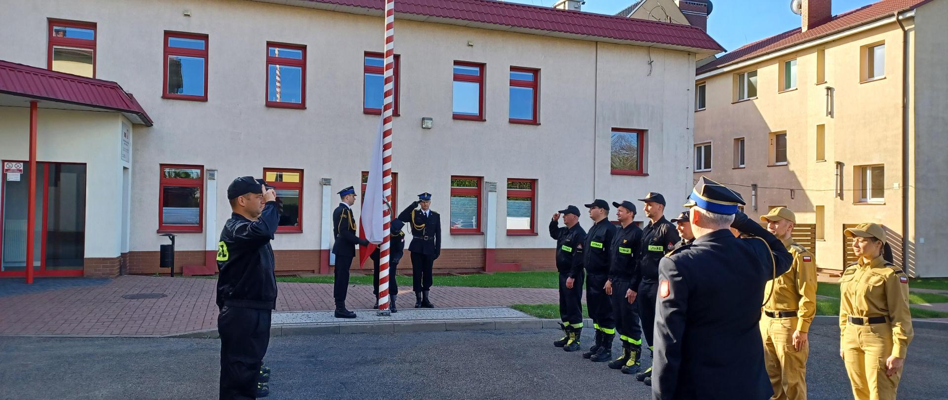 Uroczystość z okazji Dnia Flagi Rzeczypospolitej Polskiej. Strażacy ze zmian służbowych i funkcjonariusze z komendy stoją naprzeciwko siebie. Komendant, dowódcy zmian i asystujący pocztu flagowego salutują.