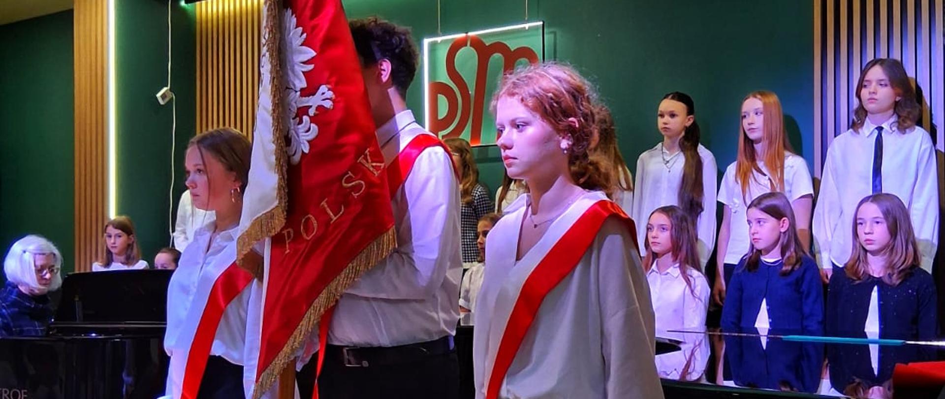 Poczet sztandarowy na scenie w sali koncertowej PSM - troje uczniów w białych koszulach z czerwono-białymi szarfami i białymi rękawiczkami, chłopak w środku trzyma czerwony sztandar z białym orłem i napisem POLSKA. W tle chór dzieci w białych i granatowych strojach oraz fortepian Petrof.