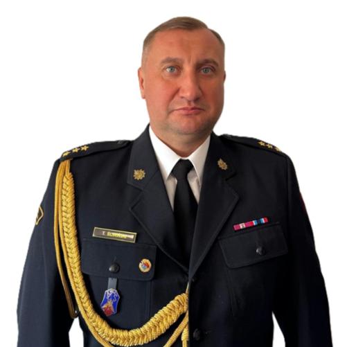 Zdjęcie przedstawia mężczyznę w formalnym mundurze służbowym. Ma krótkie, jasne włosy i poważny wyraz twarzy. Mundur jest granatowy, z krawatem, ozdobnymi złotymi sznurami na ramieniu oraz odznaczeniami i naszywkami na piersi. Tło jest jasne i jednolite.